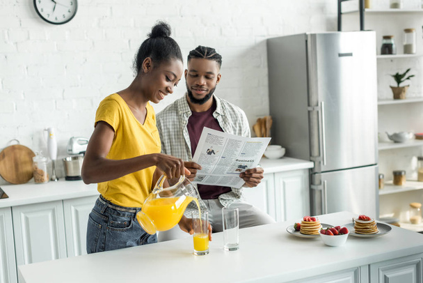 novio afroamericano leyendo periódico y novia vertiendo jugo de naranja en vidrio en la cocina
 - Foto, imagen