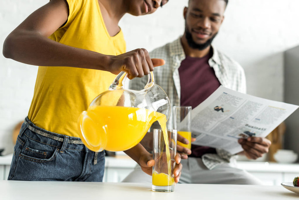 cropped image of african american boyfriend reading newspaper and girlfriend pouring orange juice in glass at kitchen - Foto, Bild