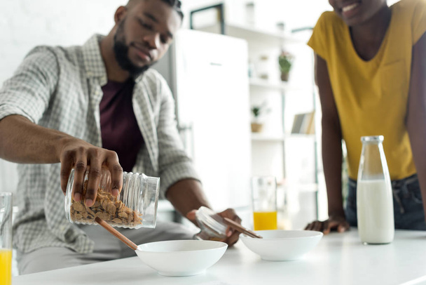 zugeschnittenes Bild eines afrikanisch-amerikanischen Freundes, der in der Küche Cornflakes in Teller gießt - Foto, Bild