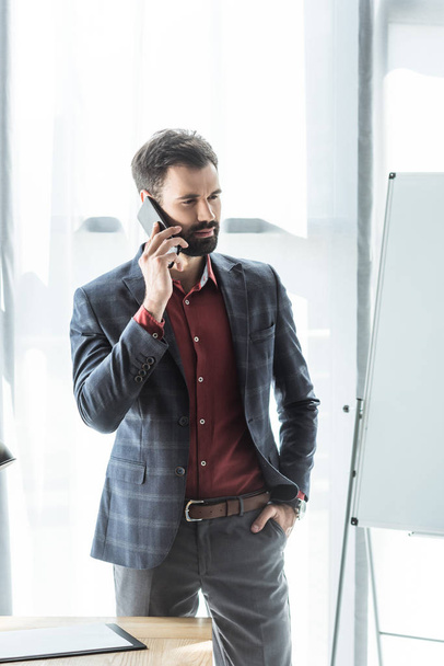 handsome young businessman in stylish suit talking by phone at office - Zdjęcie, obraz