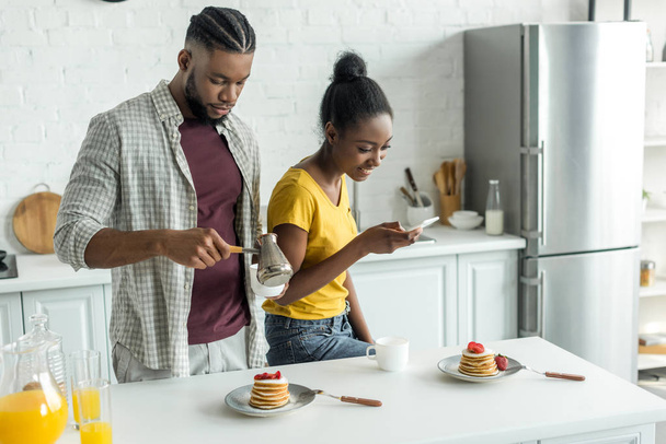 african american girlfriend taking photo of pancakes with smartphone at kitchen - Fotó, kép