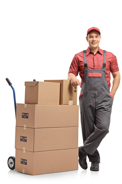 Full length portrait of a mover leaning on a stack of boxes loaded on a hand truck isolated on white background - Фото, зображення