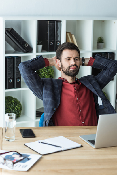 handsome young businessman relaxing at workplace with hands behind head - Foto, Imagem
