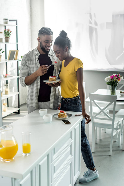 smiling african american boyfriend feeding girlfriend with strawberry at kitchen - Photo, Image