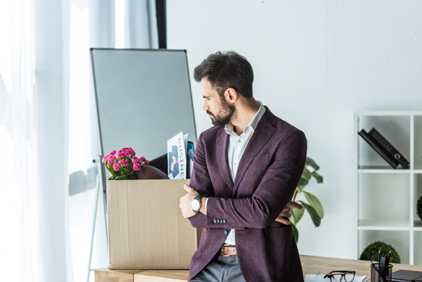 dismissed young businessman looking at box of personal stuff in office - Foto, Imagen