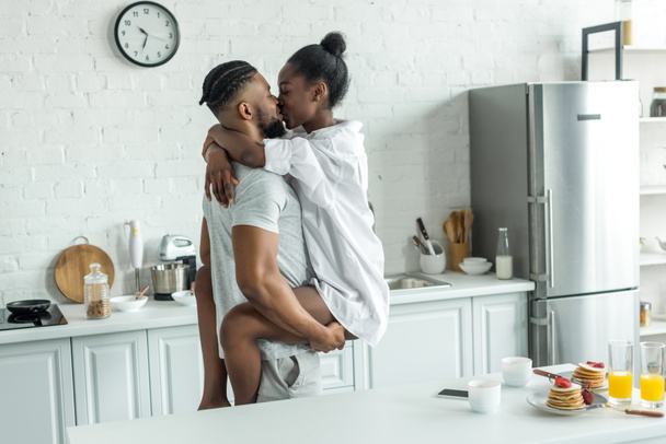 vista lateral del novio afroamericano sosteniendo novia y se besan en la cocina
 - Foto, imagen