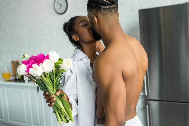 african american couple kissing and girlfriend holding bouquet at kitchen - Photo, Image