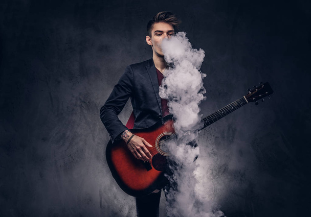 Guapo joven músico con el pelo elegante en ropa elegante exhala humo mientras toca la guitarra acústica
. - Foto, Imagen