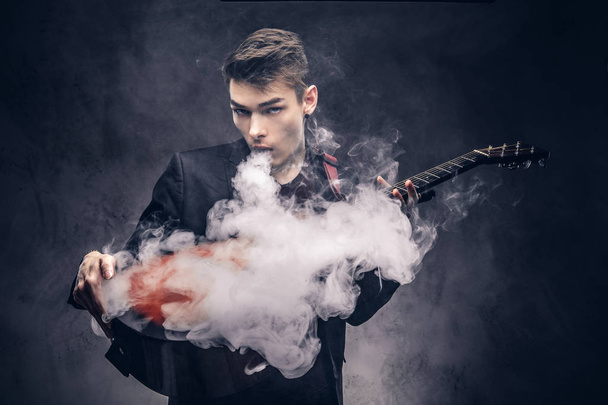 Красивый молодой музыкант со стильными волосами в элегантной одежде выдыхает дым на акустической гитаре
. - Фото, изображение