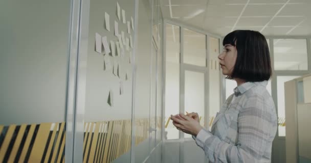 Young woman with short hair cut writing on the paper notes some ideas and stiks on the wall - Materiał filmowy, wideo