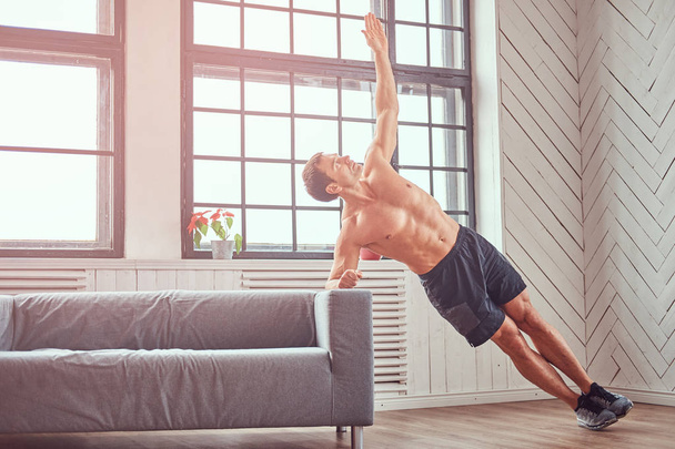 Handsome shirtless muscular male does exercise leaning on a sofa at home. - 写真・画像