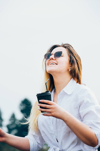uma menina de cabelos longos em óculos de sol com uma bebida, sorrindo
 - Foto, Imagem