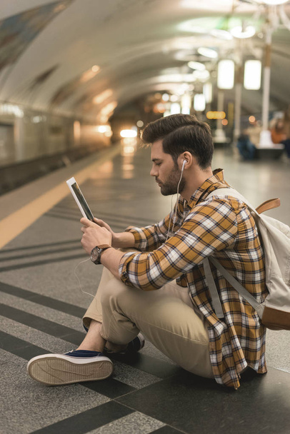 Μερική άποψη του το κομψό άνδρα με ψηφιακό tablet και ακουστικά κάθεται στο πάτωμα στο σταθμό του μετρό  - Φωτογραφία, εικόνα