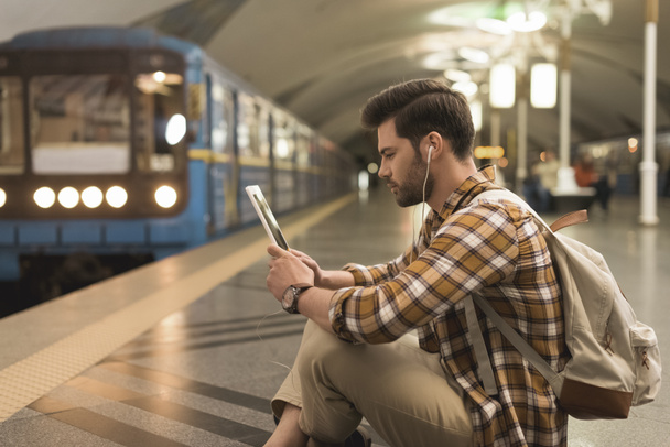 вид сбоку молодого человека с цифровым планшетом и наушниками, сидящего на полу на станции метро
  - Фото, изображение
