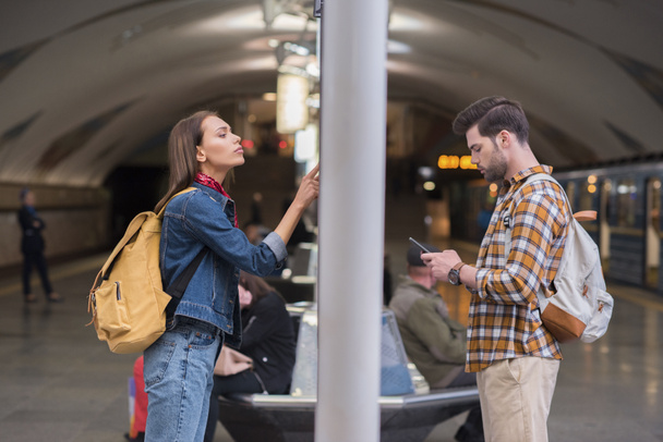 Zijaanzicht van de vrouwelijke reiziger met rugzak kijken informatiebord en vriendje gebruikend smartphone bij metrostation  - Foto, afbeelding