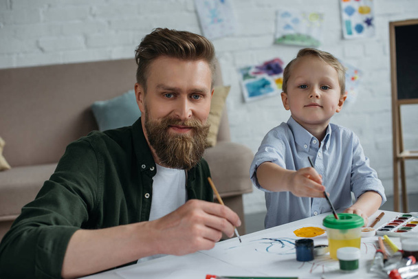 smiling father and cute little son with paints and brushes looking at camera together at home - Photo, Image