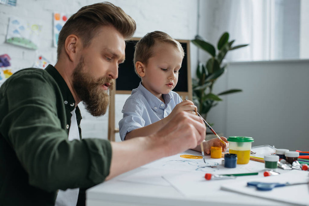 vista laterale del padre e del piccolo figlio carino con vernici e pennelli che disegnano insieme i quadri a casa
 - Foto, immagini