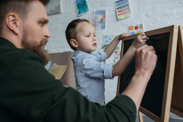 vista lateral del niño y el padre con trozos de tiza dibujo en pizarra en casa
 - Foto, Imagen