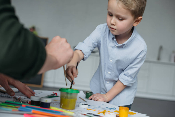 cropped shot of little boy with brush and paints drawing picture together with father at home - Fotografie, Obrázek