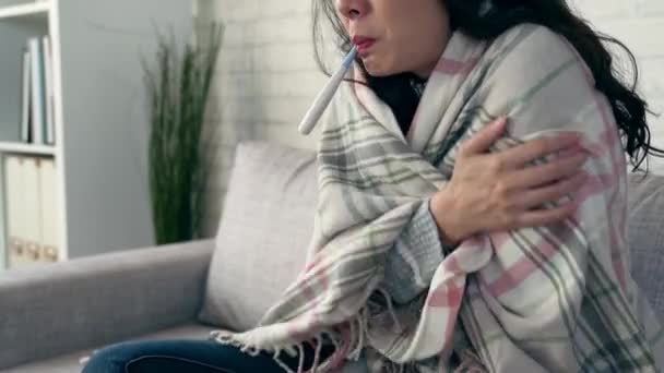 Азиатская женщина чувствует холод, и измерить температуру тела с термометром, сидя в гостиной на дому зимой
. - Кадры, видео