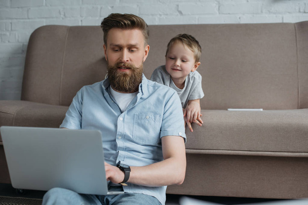 bearded man using laptop with son near by on sofa at home - Foto, afbeelding