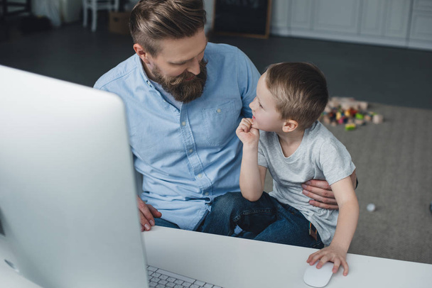 father and little son sitting at table with computer screen at home - Photo, Image