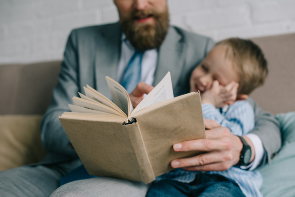selective focus of businessman and little son reading book together an sofa at home, work and life balance - Photo, Image