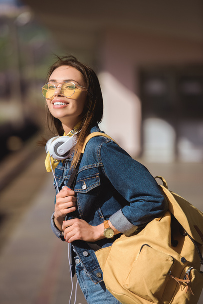 mujer elegante en gafas de sol con mochila y auriculares de pie en la estación de metro al aire libre
  - Foto, imagen