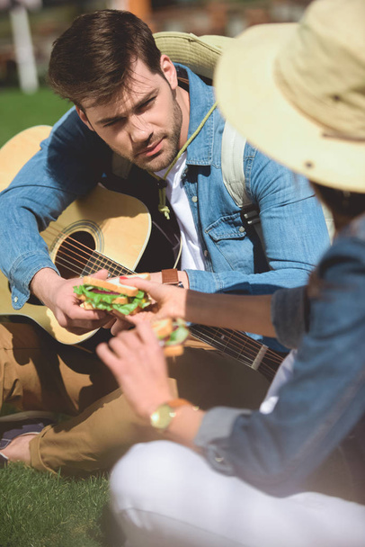 rear view of stylish female tourist giving sandwich to boyfriend with guitar  - Photo, image