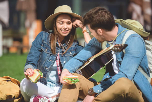 stylish travelers in hats with backpacks and guitar holding sandwiches  - Foto, Imagem
