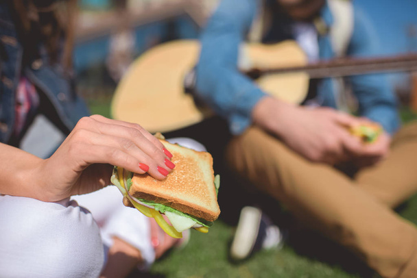 close-up tiro de sanduíche na mão feminina e homem sentado com guitarra acústica
  - Foto, Imagem