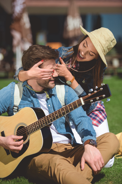 smiling female traveler in hat covering eyes of boyfriend with guitar from behind  - Φωτογραφία, εικόνα