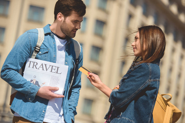 stylish female tourist giving credit card to boyfriend with travel newspaper in hand - Foto, afbeelding