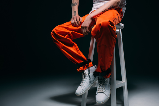 Cropped view of man in prison uniform with cuffs on legs sitting on stool on dark background - Foto, imagen