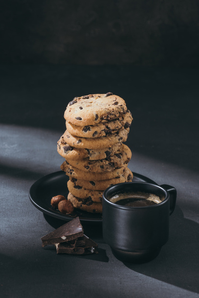 stack of chocolate-chip cookies on plate with cup of coffee on black surface - Photo, Image