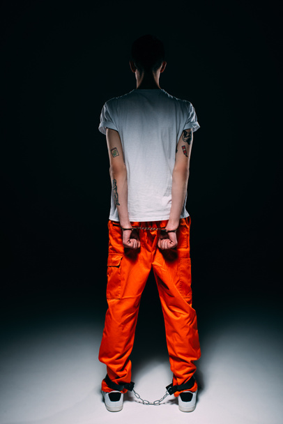Вид сзади на человека в тюремной форме с руками в наручниках за спиной на темном фоне
 - Фото, изображение