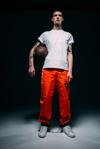 Young male prisoner wearing orange pants and cuffs holding basketball ball on dark background - Fotoğraf, Görsel