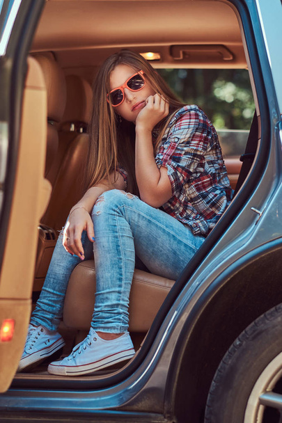 Portrait of a beautiful girl in a fleece shirt and jeans, sitting in the car in the back seat with an open door. - Photo, Image
