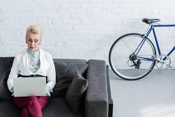 anziana donna d'affari che utilizza il computer portatile sul divano in ufficio con bicicletta
 - Foto, immagini