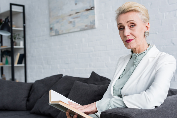 elegant senior woman reading book and sitting on sofa - Photo, image