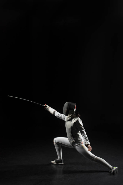 full length of female fencer in uniform posing with sword on black background - Фото, изображение