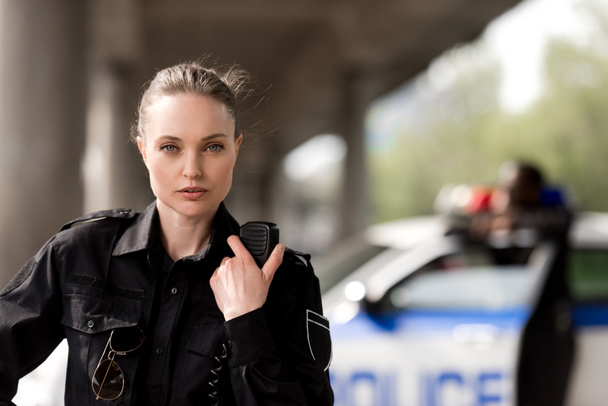 policewoman using walkie-talkie and looking at camera with blurred partner near car on background - Photo, Image