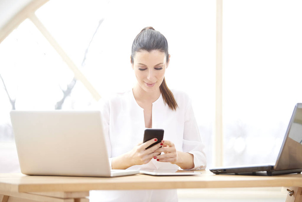Portrait of beautiful young woman using her mobile phone and text messaging while sitting at office desk in front of laptops. Smiling businesswoman managing her business from home.  - Foto, afbeelding