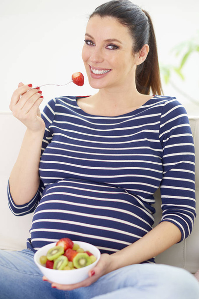 Young pregnant woman sitting on couch and eating a bowl of fruit salad.  - Φωτογραφία, εικόνα