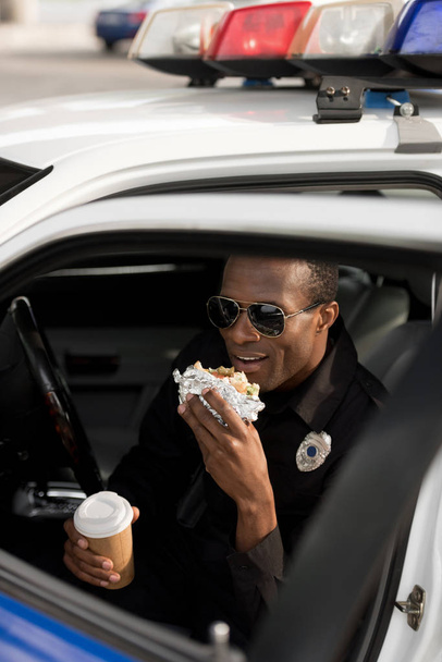 νεαρός Αφρικανός Αμερικανός αστυνομικός με φλιτζάνι καφέ, κάθονται στο αυτοκίνητο και να τρώει burger - Φωτογραφία, εικόνα
