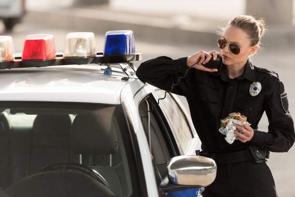 serious policewoman with burger in hand talking on radio set  - Foto, immagini