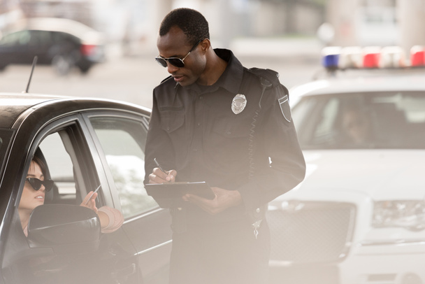 молодая женщина в машине дает водительские права африканскому американскому полицейскому с планшетом и ручкой
 - Фото, изображение