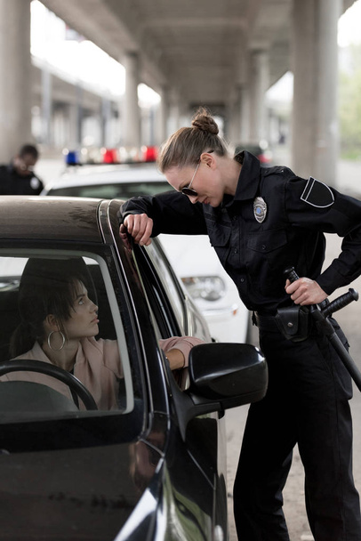 Seitenansicht einer Polizistin, die Knüppel hält und mit einer jungen Frau spricht, die im Auto sitzt  - Foto, Bild