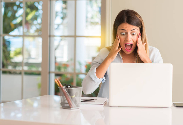 Student kobieta przy stole z laptopa w domu stresujące trzymania rąk na głowie, przerażony w panice, krzycząc - Zdjęcie, obraz