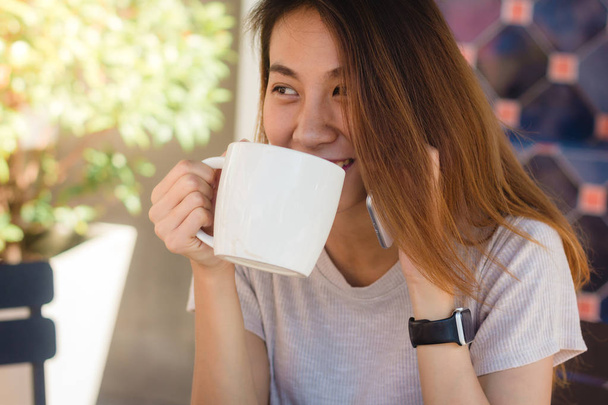 Onnellinen hymy aasialainen liike naiset käyttävät puhuva matkapuhelin istuu kahvilassa ja pitämällä kuppi kahvia. Kaunis iloinen aasialainen nainen kahvilassa puhuu puhelimessa ja hymyilee nauttien kahvista
. - Valokuva, kuva